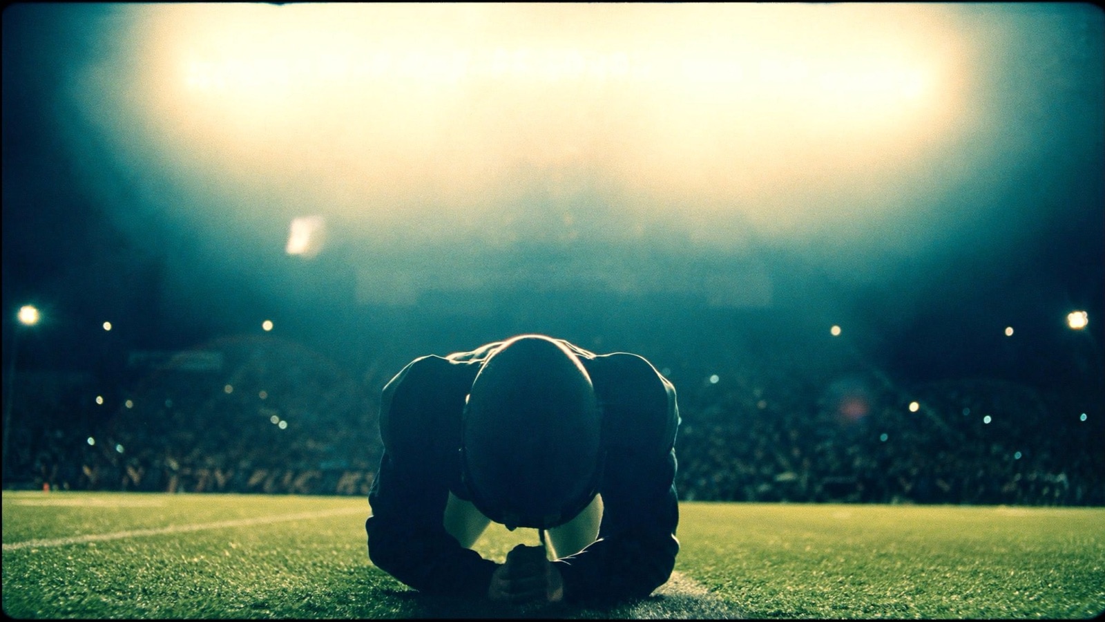 A lone football player kneeling in silhouette on the sideline under stadium floodlights.