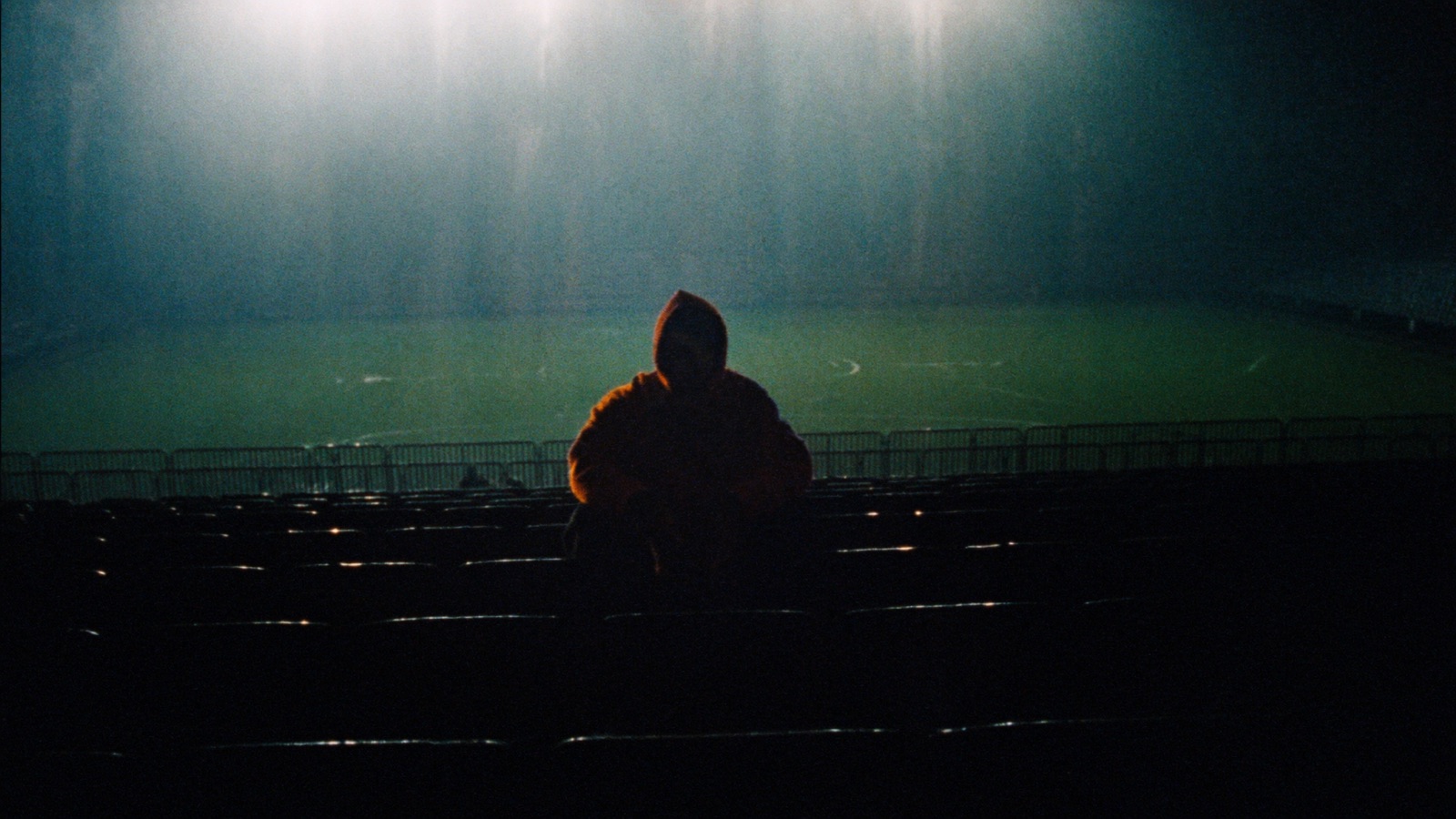 A lone figure silhouetted in empty stadium seats under floodlights, head bowed, waiting.