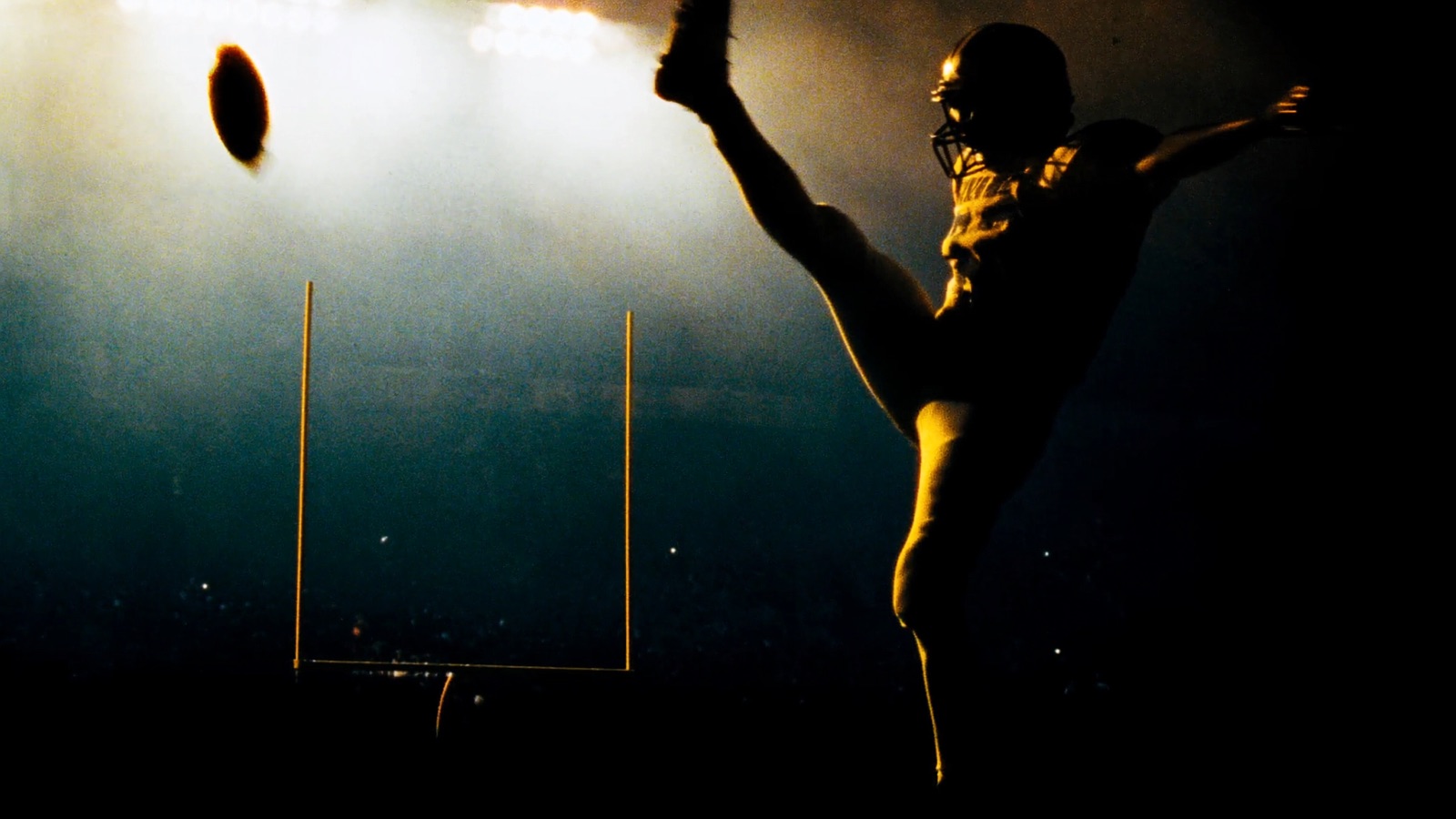 A placekicker silhouetted at the end of his follow-through as the ball sails wide of an upright under floodlights.