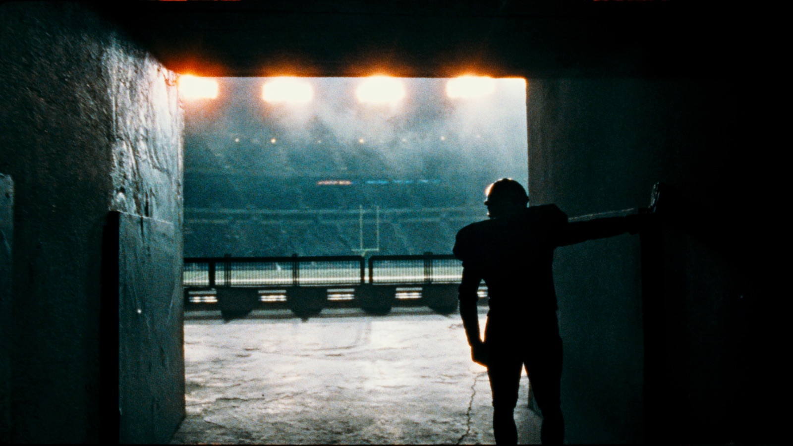 A lone football player silhouetted at the mouth of a stadium tunnel, waiting to walk out onto an empty floodlit field.
