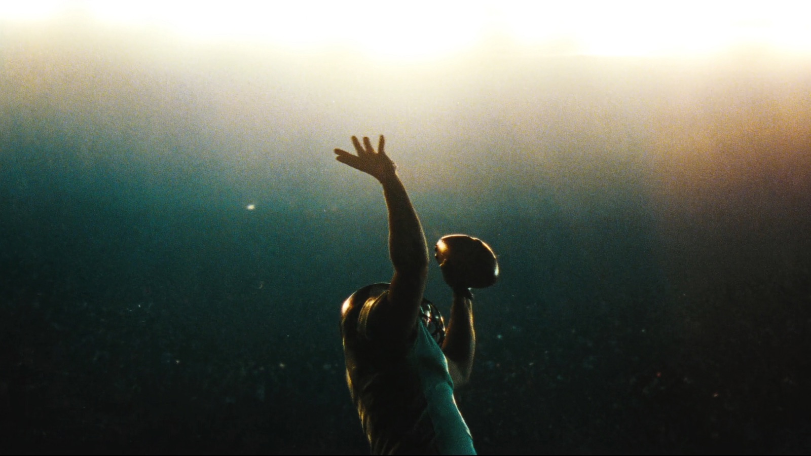 A quarterback silhouetted at the top of his throwing motion under stadium floodlights, the ball still in his raised hand.